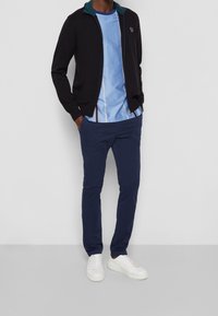 Hombre vestido con chaqueta de cremallera azul marino, camiseta a rayas azul claro, pantalones azul marino y zapatillas blancas, de pie con las manos en los bolsillos contra una pared blanca.