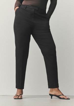Personne portant un pantalon noir ajusté et des sandales noires à talons, debout les mains dans les poches devant un fond uni.