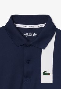 Granatowa koszulka polo z teksturowanej tkaniny, z białym paskiem po lewej stronie, trzema guzikami i zielonym logo krokodyla na pasku.