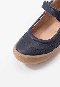 Navy kožená bota s kulatou špičkou, zdobená dekorativními perforacemi a vlnitým vzorem, zajištěná páskem přes nárt. Gumová podrážka.