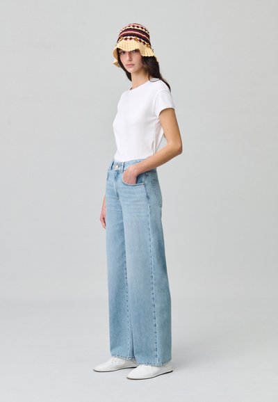 T-shirt en coton blanc associé à un jean en denim large bleu clair. Le mannequin porte un chapeau seau en crochet avec un motif jaune et bordeaux. Chaussures blanches.