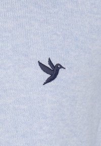 Ljusblått stickat tyg med en mjuk textur som har ett litet mörkblått broderat kolibri-emblem på vänster sida.