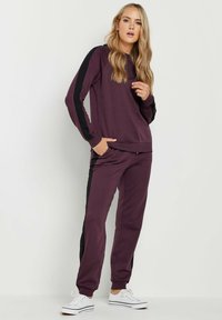 Lila Sweatshirt und passende Hose mit schwarzen Seitenstreifen. Hergestellt aus weichem Material, mit lockerer Passform und Fronttaschen. Weiße Sneaker.