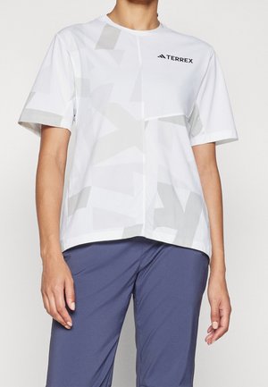 Osoba ubrana w luźny biały krótki T-shirt TERREX z subtelnymi geometrycznymi wzorami oraz niebieskie spodnie, pokazana od ramion do ud.