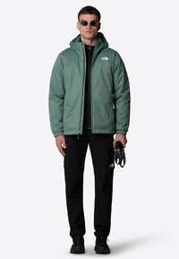 Grøn zip-up jakke med hætte, sort basislag, sorte cargo bukser og sorte vandrestøvler; holder handsker. Har et hvidt logo.