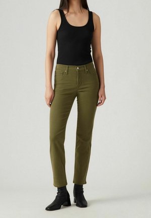 Femme portant un haut noir sans manches, un jean droit vert olive et des bottines noires à talons, debout devant un fond uni.