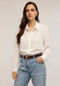 Blouse blanche à manches longues avec un devant à boutons et un col, assortie à un jean en denim bleu clair et une ceinture marron foncé avec une boucle en or.