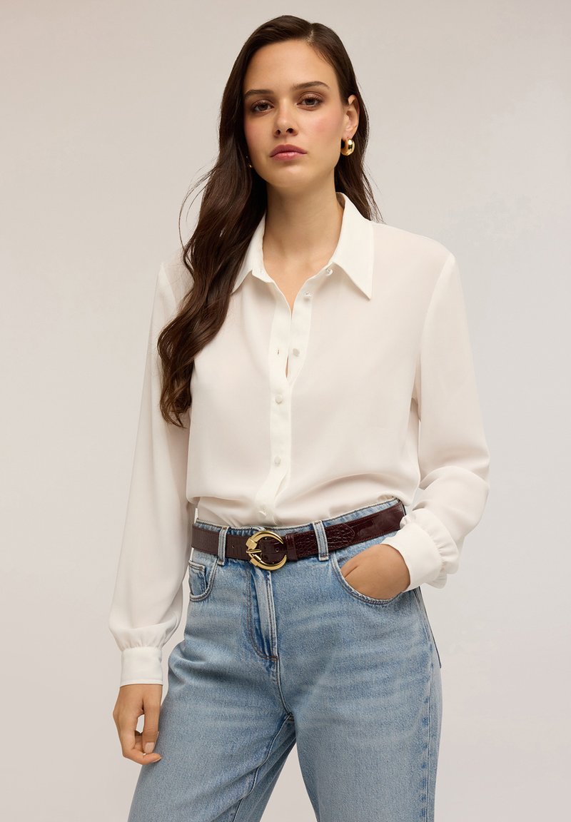 Blouse blanche à manches longues avec un devant à boutons et un col, assortie à un jean en denim bleu clair et une ceinture marron foncé avec une boucle en or.
