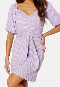 Robe à carreaux lilas avec des manches bouffantes, un décolleté en V profond et un détail de taille nouée. Fabriquée en tissu léger avec une finition texturée.