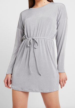 Kobieta ubrana w jasnoszarą mini sukienkę z długim rękawem, okrągłym dekoltem i wiązanym paskiem w talii.