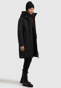 Fekete parka kapucnival, elülső gombokkal, oldalsó zsebekkel és cipzáras záródással. Fekete leggings-szel és vaskos fekete csizmával párosítva. Sima textúra.