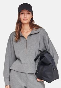 Graue Sweatjacke mit Reißverschluss, locker sitzend und mit einem Volant-Saum, kombiniert mit einer schwarzen Kappe und einer schwarzen Tasche. Glatte Stofftextur.