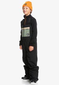 Svart snöbyxa med justerbara axelremmar, med en grön bröstficka och en beige klaff. Modellen bär en orange mössa och svarta handskar.
