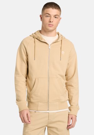 Joven con una sudadera beige con cremallera, cordones y bolsillos frontrales, combinada con pantalones beige a juego, sobre un fondo blanco.