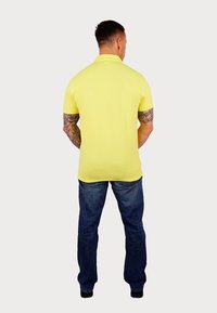 Sárga póló, rövid ujjú, slim fit fazon, kék farmernadrággal párosítva, hátulról nézve, látható tetoválásokkal a karokon.
