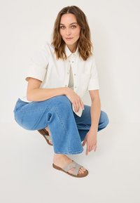 White collared shirt, blue wide-leg jeans, and brown sandals with cross-strap design. Model crouches in a minimalist setting.