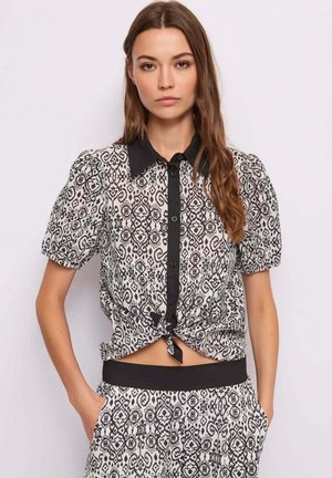 Giovane donna che indossa una camicia a maniche lunghe bianca e nera con bottoni, annodata in vita, e pantaloni coordinati con motivo e cintura nera.