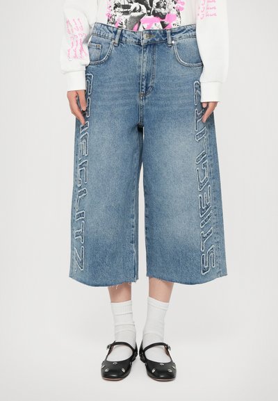 Hellblaue Denim-Culottes mit ausgefransten Kanten, versehen mit dem gestickten Schriftzug "STIEGLITZ" an der Seite. Kombiniert mit schwarzen Clogs und weißen Socken.