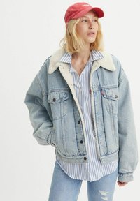 Lichtblauwe spijkerjack met een crèmekleurige sherpa-kraag, voorzien van twee voorkezen en een relaxed fit. Draagt over een blauw-wit gestreept shirt.