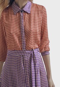 Vestido con patrón de pata de gallo en bloques de color rojo y púrpura, con cuello de solapa y un cinturón atado en la cintura para dar forma definida.