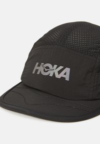 Casquette noire avec panneaux en mesh, logo "HOKA" réfléchissant à l'avant, visière courbée, tissu texturé et détails de couture sur la visière.