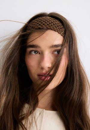 Young woman with long brown hair wearing a brown knitted headband, looking slightly to the side with hair gently blowing across her face.