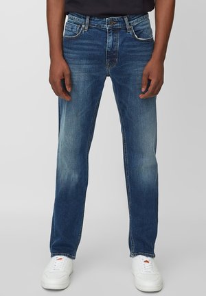 Homme portant un jean bleu délavé droit et des baskets blanches, debout devant un fond clair uni.