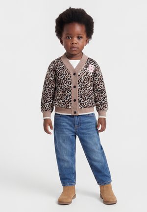 Bambino piccolo in piedi, indossa un cardigan leopardato con la lettera "G", jeans blu e stivali color sabbia, su uno sfondo bianco semplice.