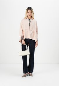 Blusa rosa con bottoni e orlo a peplo, indossata sopra una camicia a maniche lunghe con fantasia. Pantaloni flare in denim scuro e una borsa bianca completano il look.