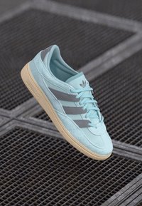 Světle modré semišové tenisky se třemi šedými pruhy, gumovou podrážkou, texturovaným svrškem a jemným brandingem. Šnúrky jsou sladěny s barvou boty.