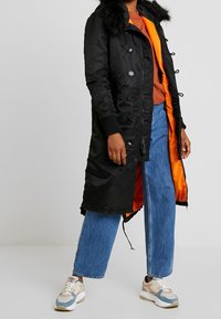 Czarna, ocieplana kurtka z kapturem wykończonym sztucznym futrem, pomarańczową podszewką, zatrzaskami i sznurkiem w talii, zestawiona z niebieskimi jeansami o szerokich nogawkach i sneakersami.
