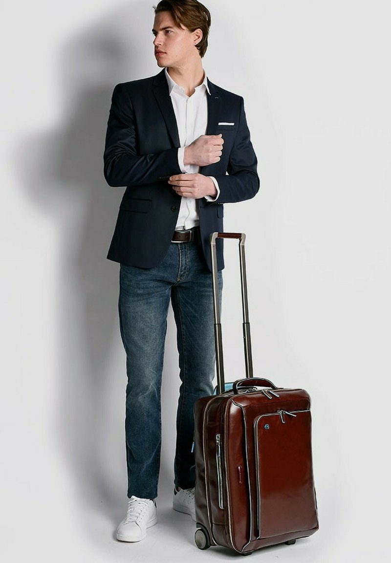 Brauner Lederkoffer auf Rollen mit doppeltem Reißverschluss, Seitentasche und ausziehbarem Griff. Kombiniert mit einem marineblauen Blazer und blauen Jeans.