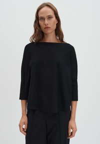 Femme aux cheveux bruns ondulés portant un haut noir ample à manches trois-quarts et un pantalon noir, devant un fond gris clair uni.