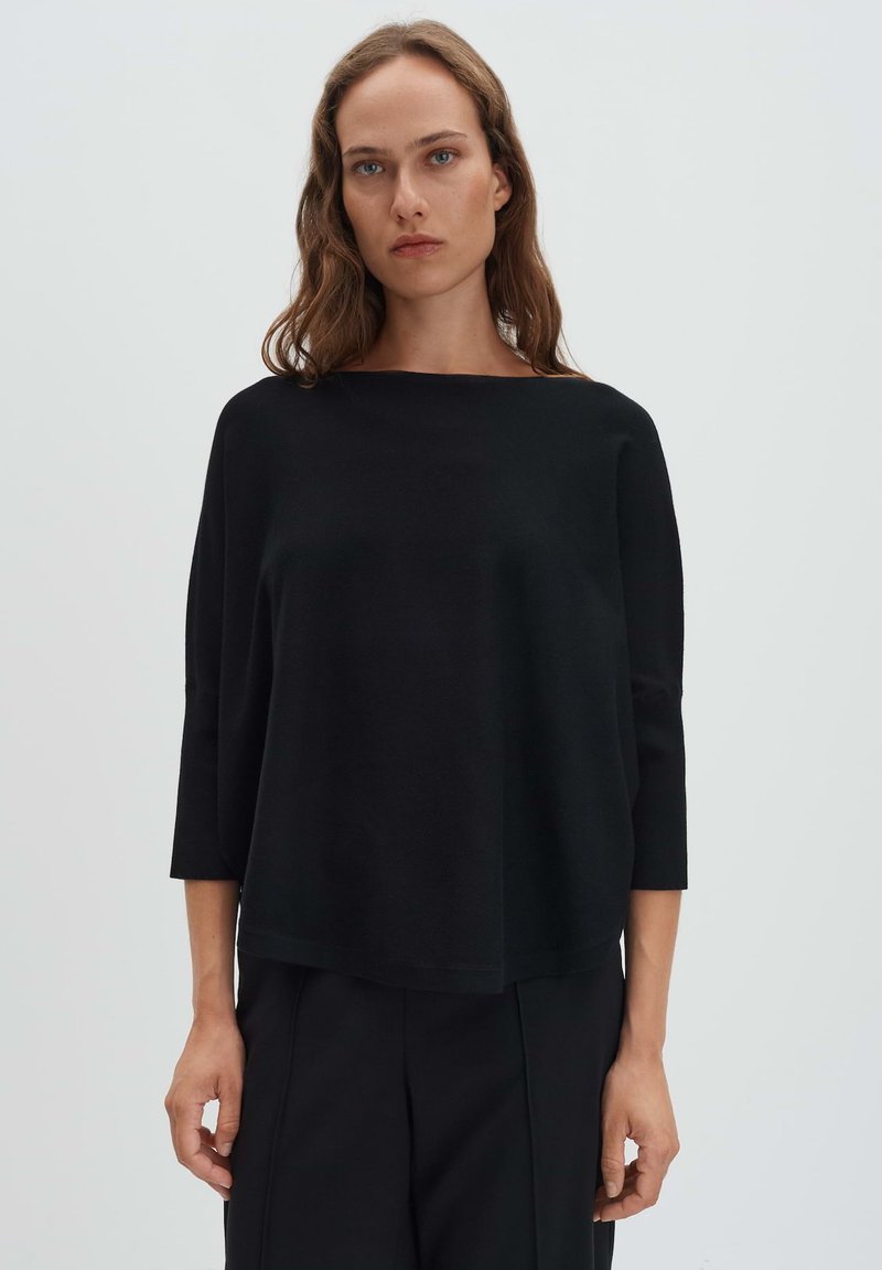 Femme aux cheveux bruns ondulés portant un haut noir ample à manches trois-quarts et un pantalon noir, devant un fond gris clair uni.