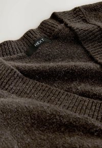 Mørkebrun strikket sweater med ribkant ved kraven og ærmekanterne. Har en tekstureret, blød stof. Mærke synligt med brandnavnet "NEXT."