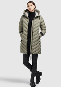 Ljust olivgrön puffjacka med avtagbar huva, diagonal quiltat mönster, framdragkedja och två sidofickor. Matchad med svarta leggings och stövlar.