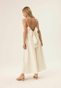 Weißes, ärmelloses Maxikleid mit einem tiefen V-Rücken und einem Rüschendetail. Der Stoff wirkt weich mit einer fließenden, glatten Textur.