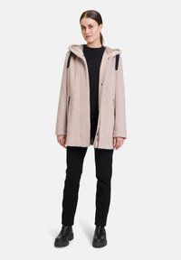 Cappotto beige chiaro con texture e cappuccio, caratterizzato da dettagli neri e tasche laterali, abbinato a pantaloni neri e stivali neri ingombranti.