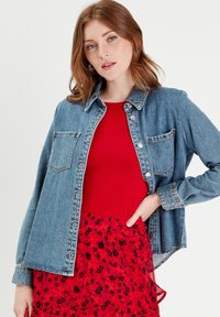 Camicia di denim azzurro chiaro con maniche lunghe, due tasche sul petto e bottoni argentati. Abbinata a un top rosso a coste e stratificata con una gonna floreale rossa.