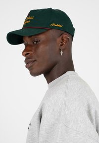 Groene baseballpet met geborduurde tekst en kastanjebruine accenten. Model draagt een grijze sweatshirt met een textuurafwerking en een kruisoorbel.