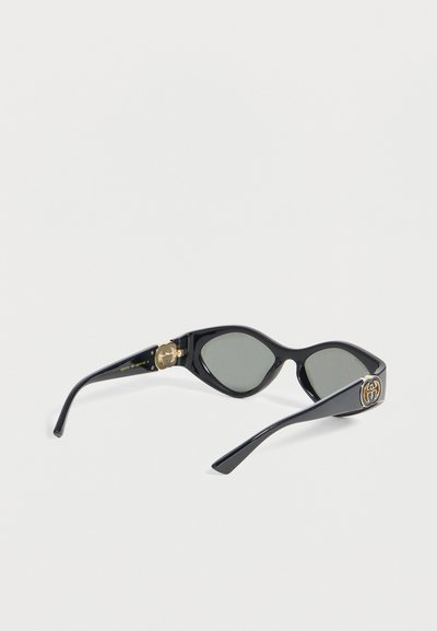 Μαύρα γυαλιά ηλίου σε σχήμα γατίσιου ματιού με σκούρους φακούς και χρυσά λογότυπα Gucci στους δύο βραχίονες, εμφανιζόμενα σε λευκό φόντο.