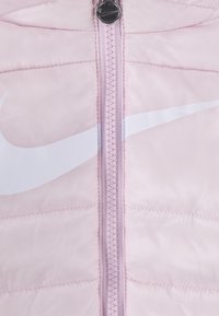 Gaiši rozā pūkainā jaka ar priekšējo rāvējslēdzēju, gaiši rozā izšuvumiem un lielu baltu Nike logotipu kreisajā pusē.