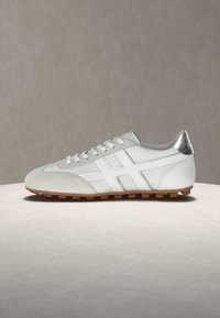 Άσπρο sneaker χαμηλού ύψους με γκρι λεπτομέρειες, κλείσιμο με κορδόνια, υφή σόλας από καουτσούκ και αντανακλαστικό ασημί ταμπελάκι στη φτέρνα, παρουσιάζεται σε ουδέτερη επιφάνεια.