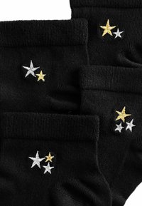 Schwarze Knöchelsocken mit gestickten Sternen in Weiß und Gold, mit gerippten Bündchen und einer weichen Textur. Drei Sterne auf einer Socke, zwei auf der anderen.