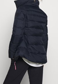 Marinblå puffjacka med hög krage, horisontell kviltad design, sidofickor och dragsko i nederkant med accentfärger.