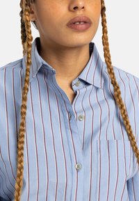 Light blue button-up shirt featuring vertical red stripes, a chest pocket, and a collar with a soft texture. Two buttons are visible.