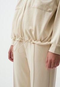 Beige ritsjack met een gathered taille, voorzien van touwtjes en twee zakken aan de voorkant. Glad materiaal met een casual silhouet.