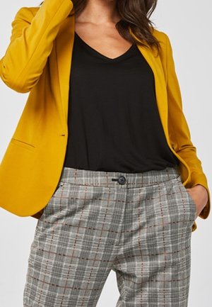 Kvinde iført en sennepsgul blazer, sort V-hals top og grå ternede højtaljede bukser med hænderne i lommerne.