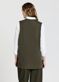 Gilet senza maniche di colore verde oliva con un design strutturato, una consistenza sottile e un taglio dritto. Abbinato a una camicia bianca e a pantaloni larghi.