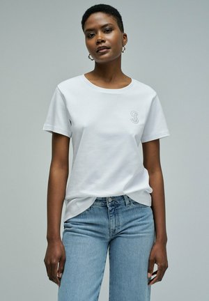 Mujer con camiseta blanca de manga corta con una pequeña letra "S" bordada y jeans azul claro, de pie contra un fondo gris liso.